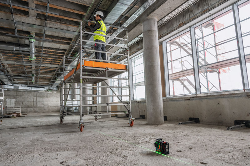 Osoba v bezpečnostním vybavení pracuje na lešení s laserovým nivelačním přístrojem na podlaze.