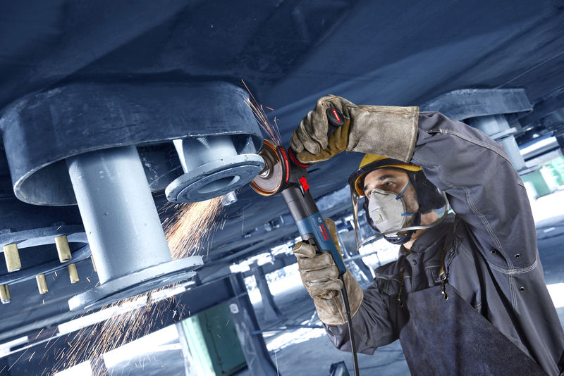 Osoba v bezpečnostním vybavení brousí kov nad hlavou úhlovou bruskou a vytváří jiskry.