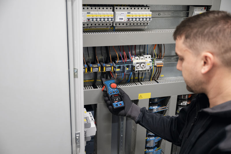 Osoba v bezpečnostním vybavení měří napětí v elektrickém rozvaděči pomocí klešťového měřiče.