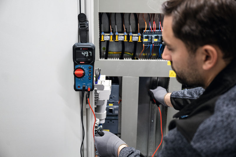 Osoba v bezpečnostním vybavení používá digitální multimetr k testování elektrického panelu.