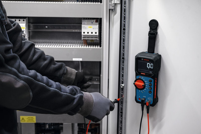 Osoba v bezpečnostním vybavení používá digitální multimetr ke kontrole elektrického rozvaděče.